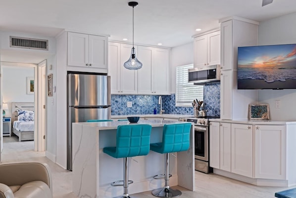 Une cuisine moderne avec des meubles blancs, un réfrigérateur en acier inoxydable, un pare-feu en carreaux bleus et un coin bar avec des tabourets turquoise.
