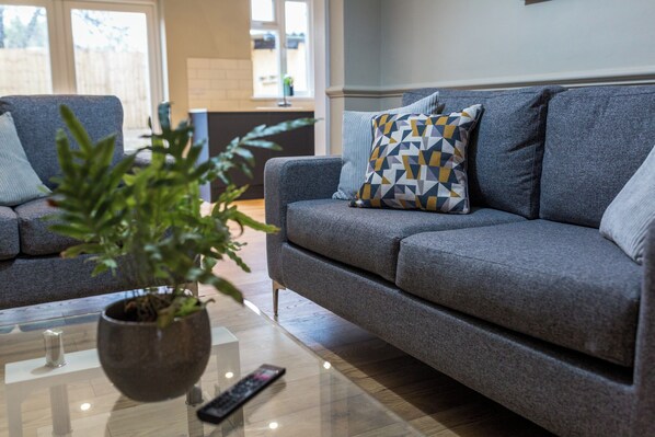 A modern living room with a grey sofa, a patterned throw pillow, a potted plant, and a remote control on a glass coffee table.