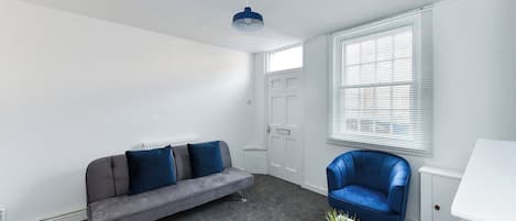 A room with a grey sofa, a blue armchair, a white table, and a window with blinds.