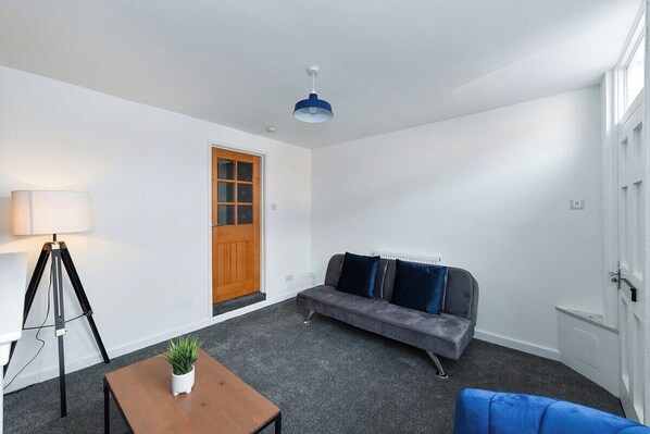Um quarto com um sofá cinzento, uma pequena mesa com uma planta, um abajur e uma porta com janela.