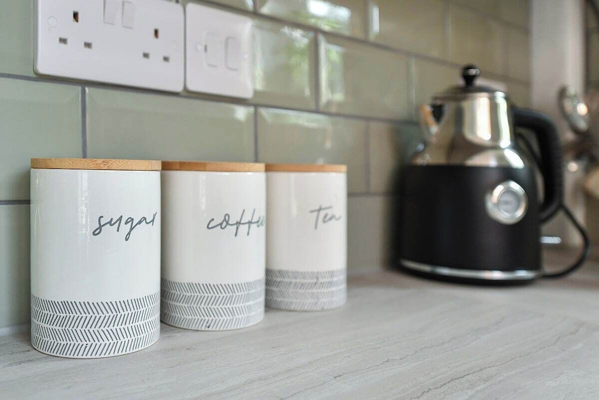 Trois récipients blancs, chacun portant une étiquette, sont posés sur le comptoir de la cuisine, à côté d’une machine à café noire et d’un mur recouvert de carreaux.
