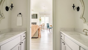 Un baño moderno con encimeras de mármol blanco, una bata color beige suave y vistas a un luminoso salón con chimenea, un sillón azul y un comedor.