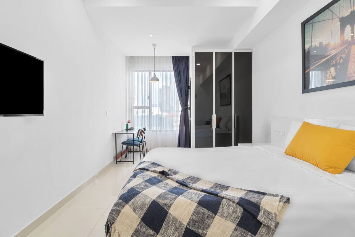 A hotel room with a bed, a desk with a chair, a window with curtains, and a painting on the wall.
