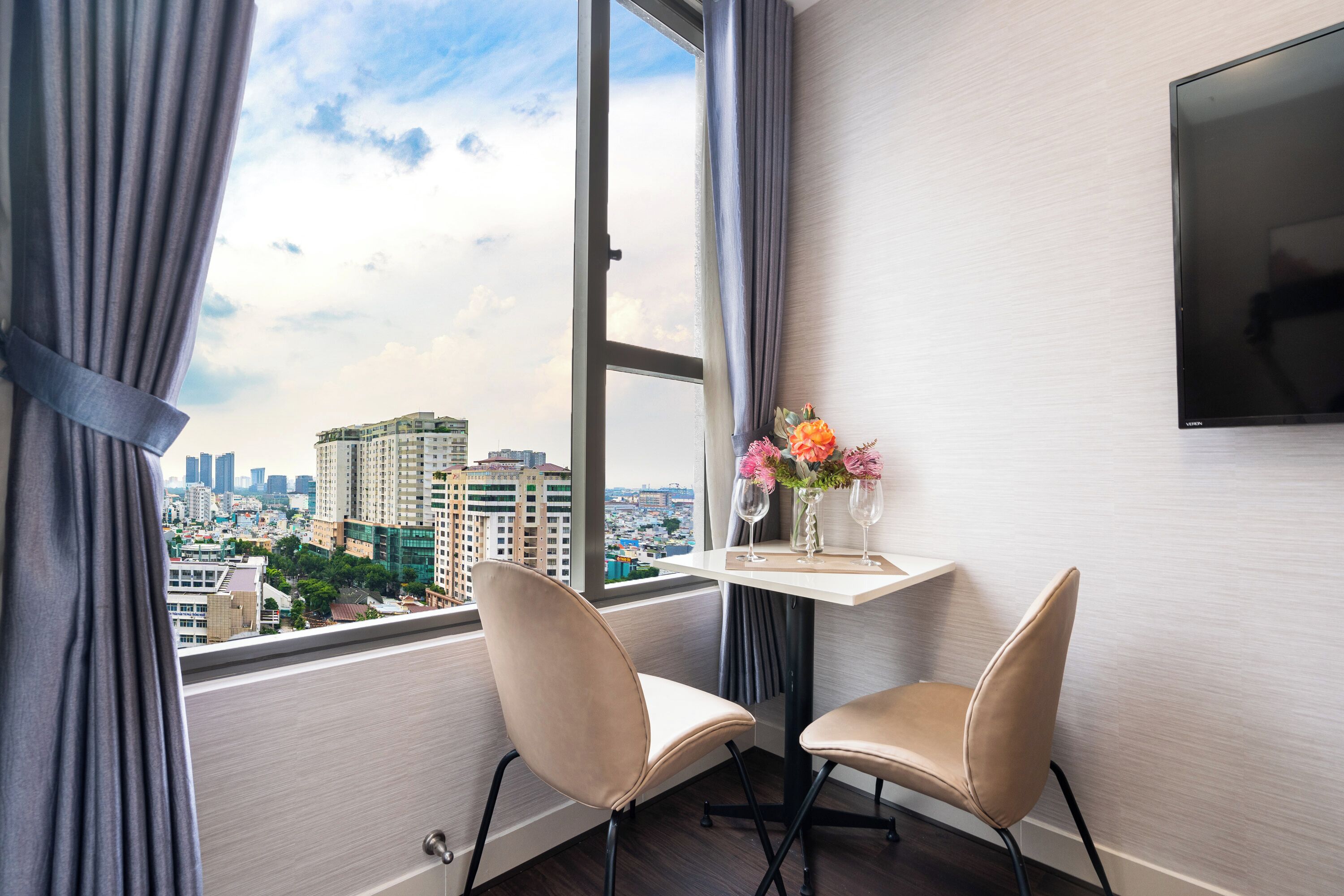 A hotel room with a large window, two chairs, a small table with a vase of flowers, and a flat-screen TV mounted on the wall.
