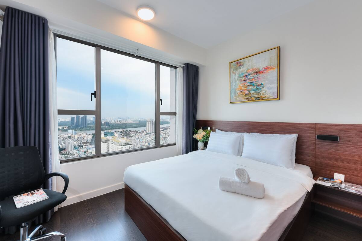 A hotel room with a large window, a bed with white linens, a chair, a nightstand, and a painting on the wall.