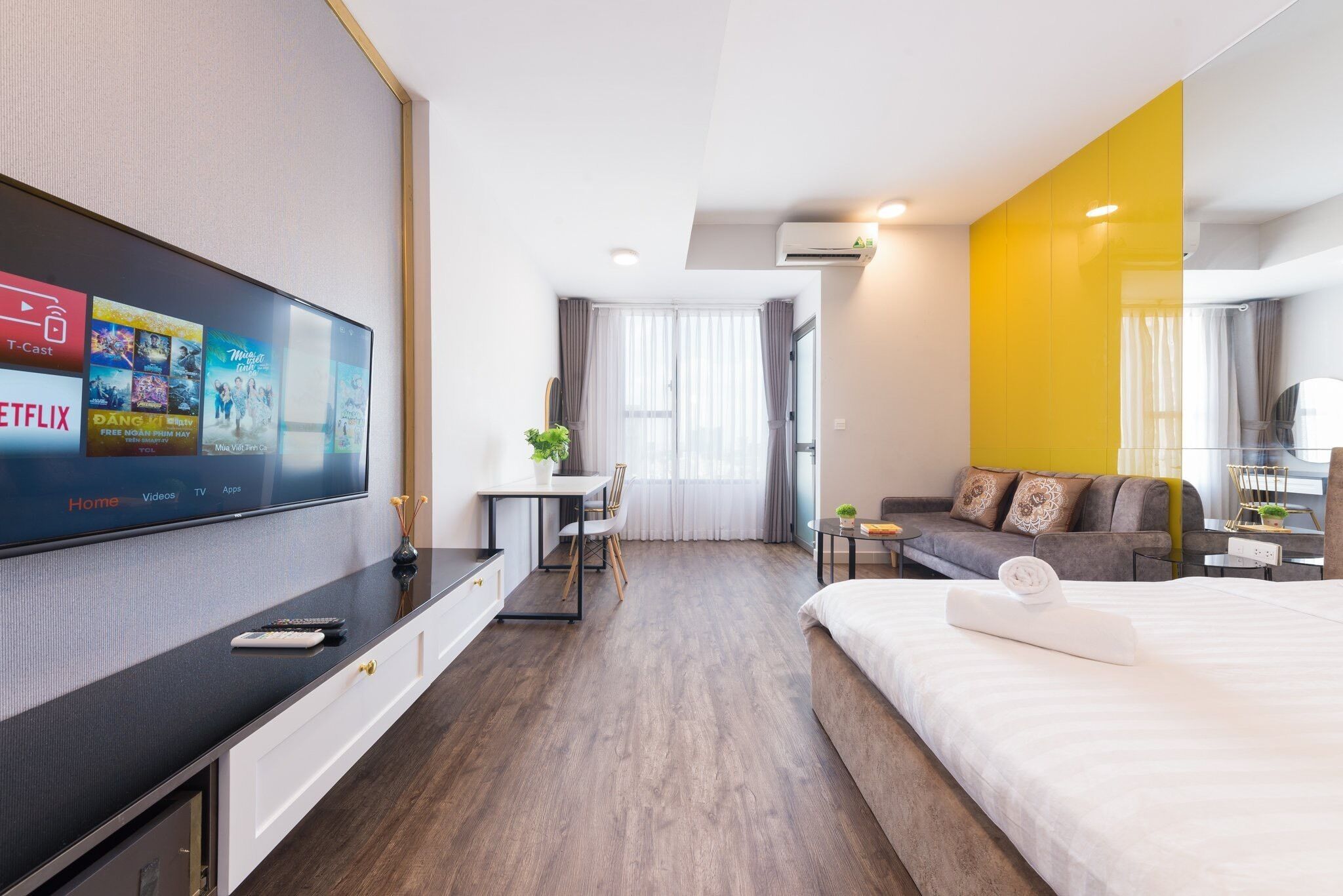 A modern hotel room with a flat-screen TV displaying a streaming service interface, a bed with a rolled towel, a sofa, a desk with a chair, and a small table with a plant.