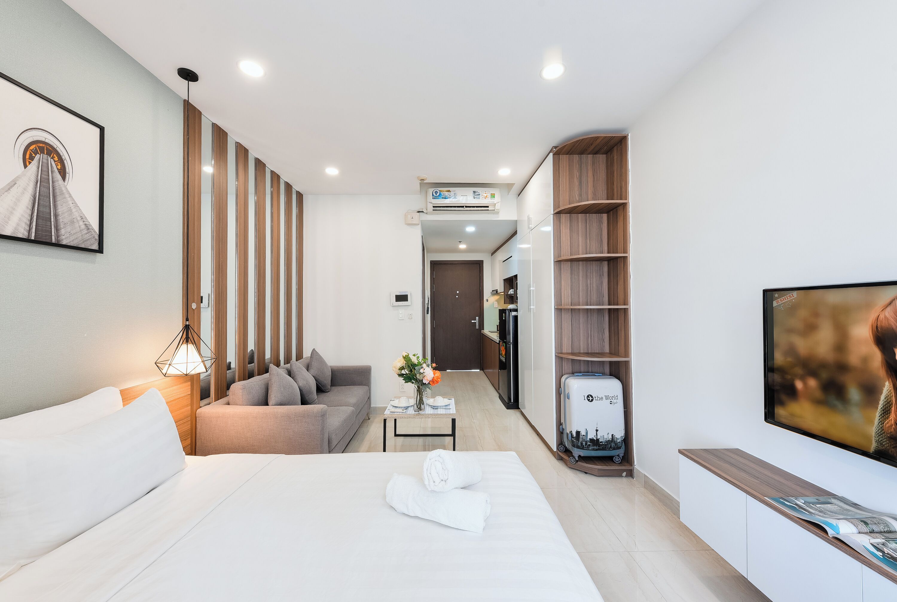 A modern hotel room with a neatly made bed, a sofa, a small table with a vase of flowers, a TV, and a bookshelf.