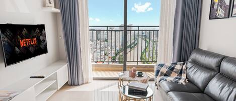 Una sala de estar moderna con un televisor de pantalla plana, un sofá de cuero, una mesita redonda con libros y velas, y un balcón con vistas a la ciudad.