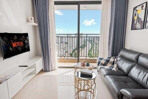 A modern living room with a flat-screen TV, a leather sofa, a small round table with books and candles, and a balcony with a city view.