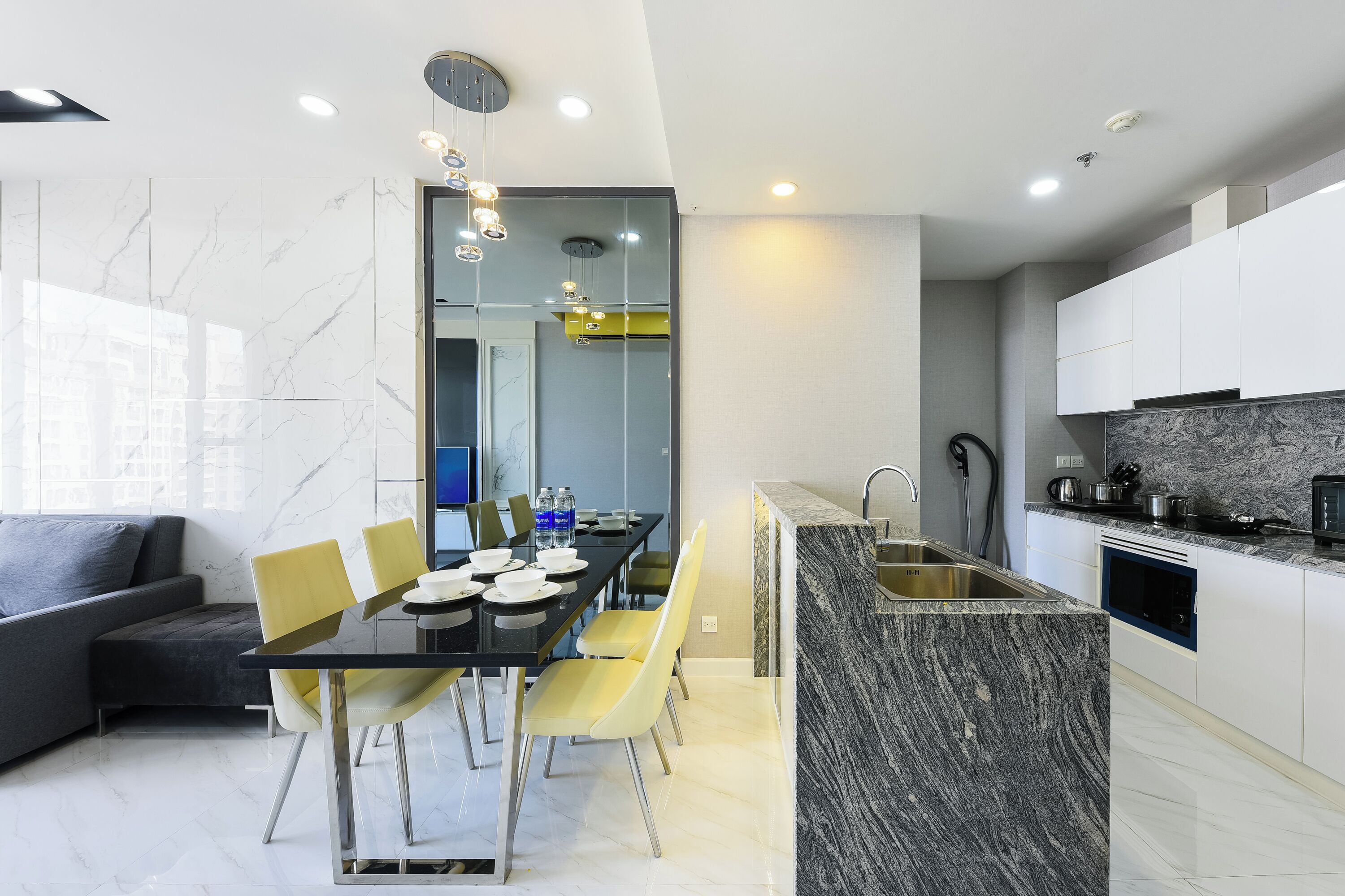 Una cocina moderna con encimera de mármol, una zona de comedor con mesa negra y sillas amarillas, y un sofá gris en la sala de estar.