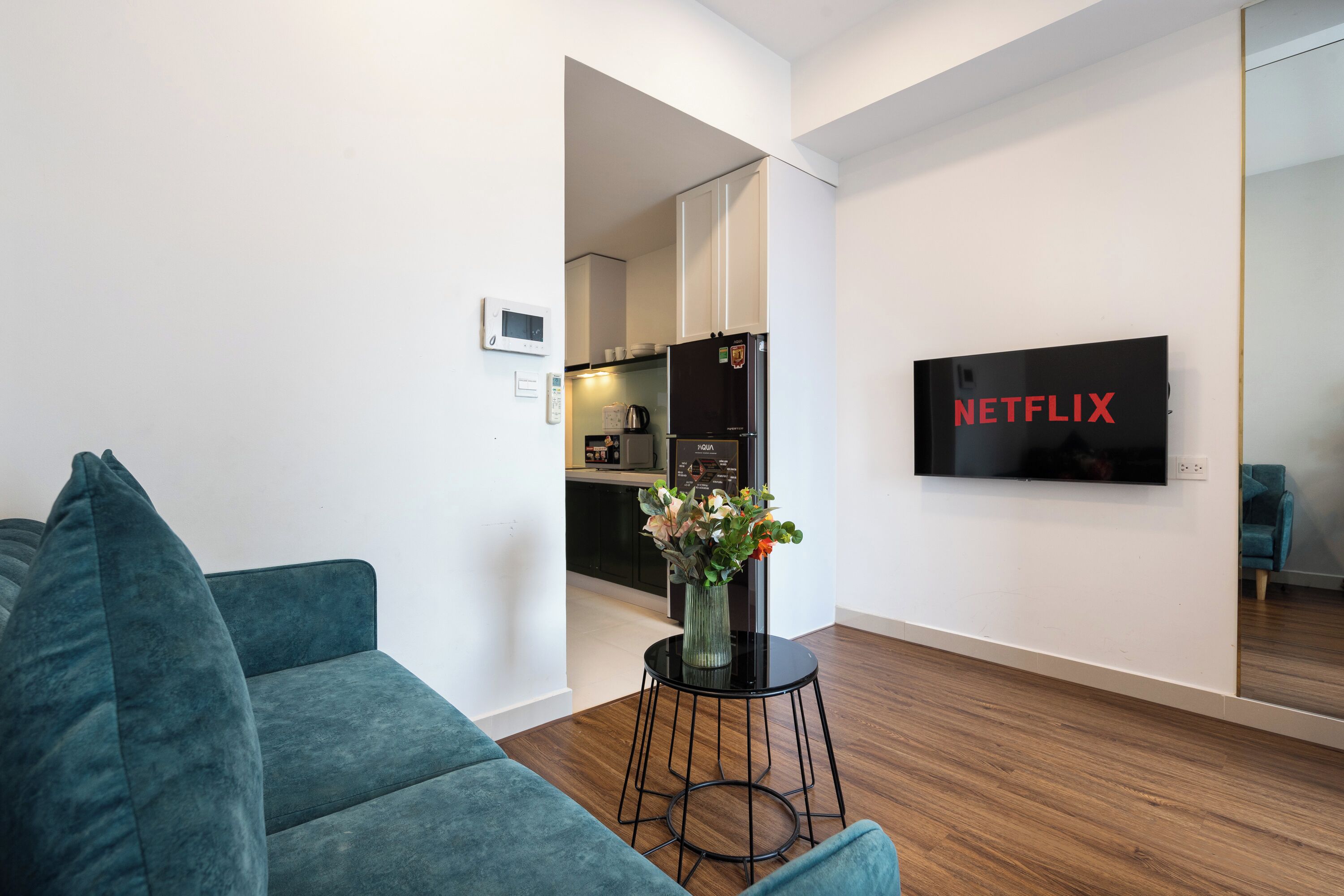 A modern living room with a teal sofa, a small black table with a vase, a kitchen in the background, and a TV displaying the Netflix logo.