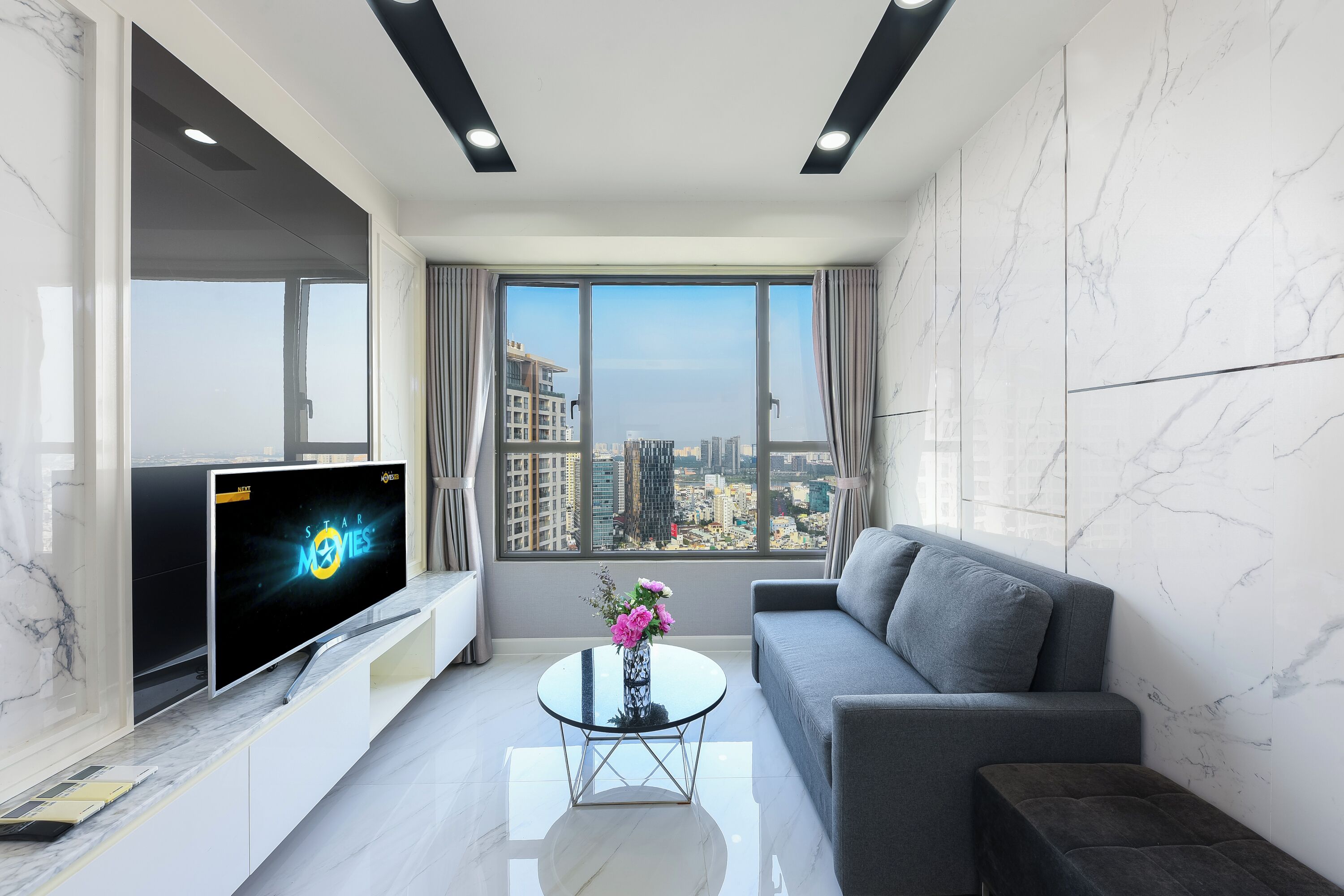Een moderne woonkamer met een grote flatscreen-tv, een bank, een salontafel met bloemen en een stadsgezicht door grote ramen.