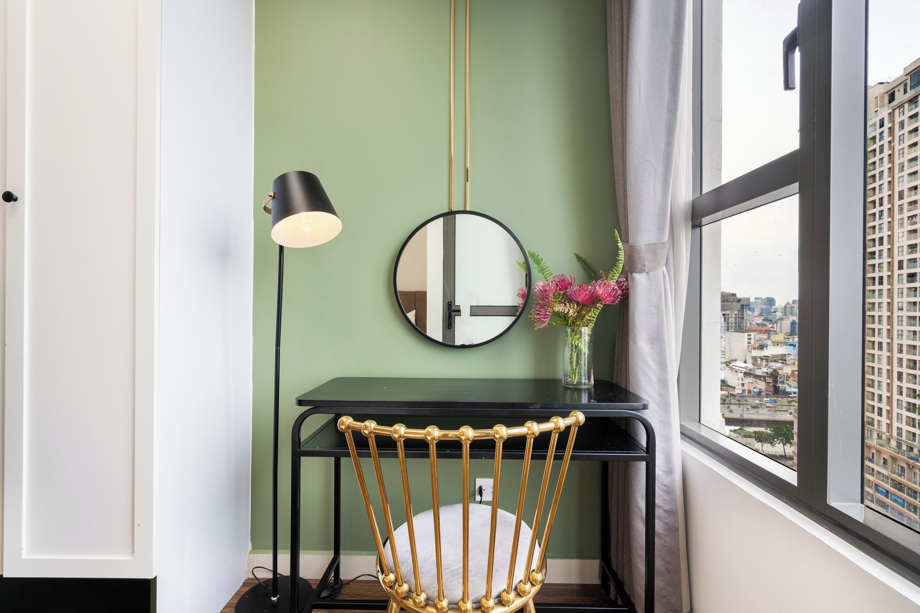 A room with a desk, a chair, a lamp, a mirror, and a window with a city view.
