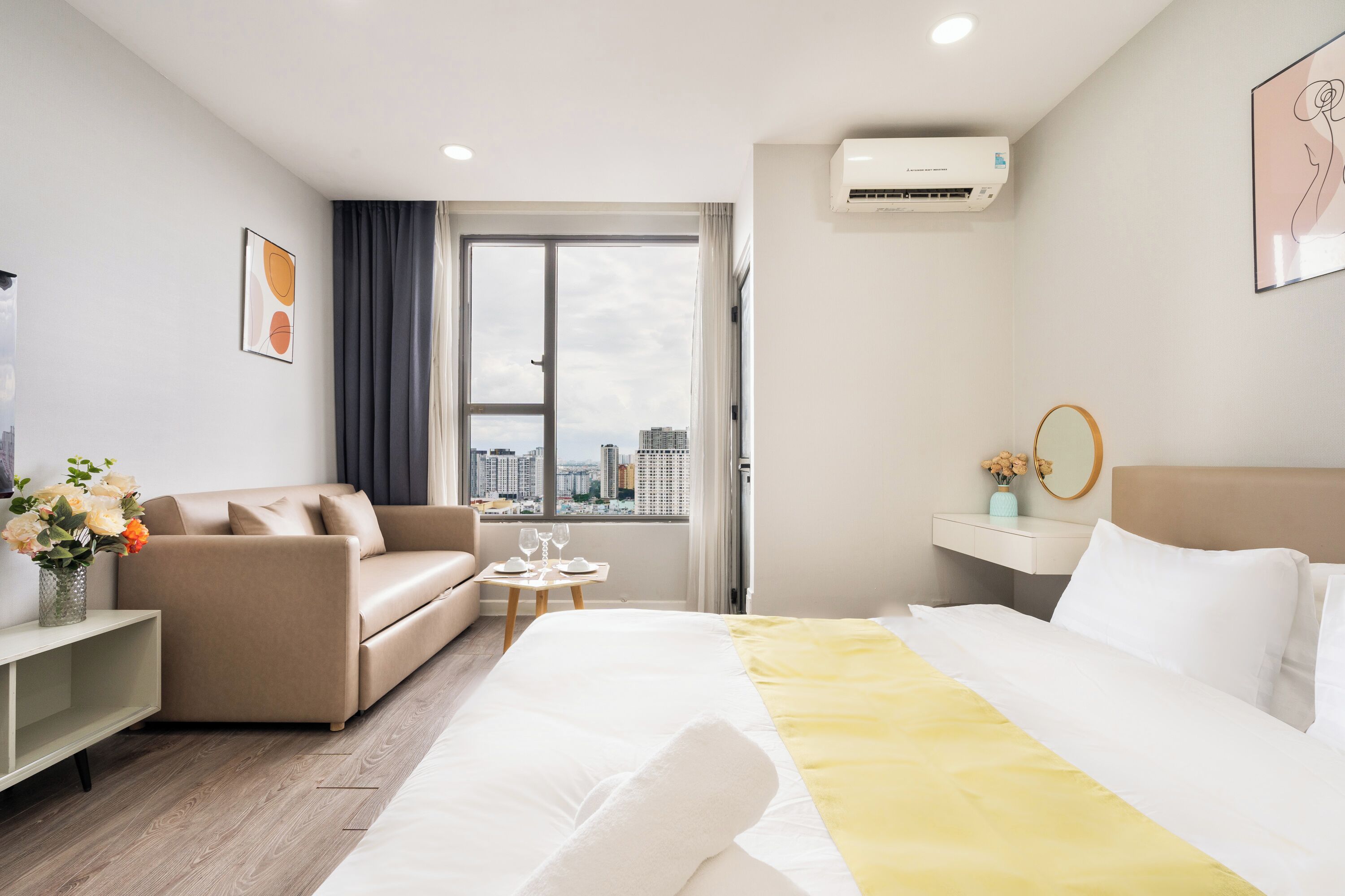 Habitación de hotel con cama, sofá, una mesita con vasos, un jarrón con flores y ventana con vistas a la ciudad.