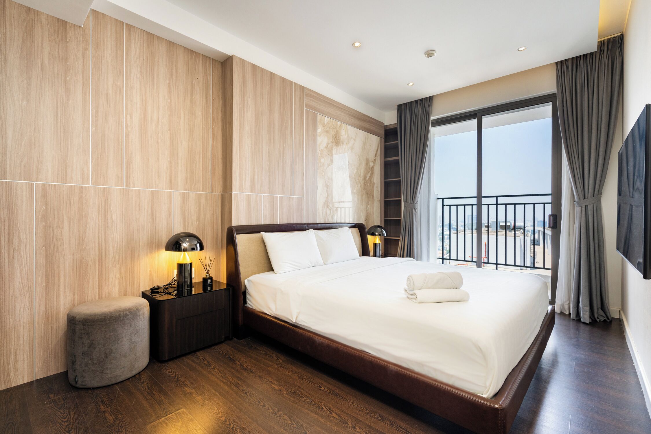 A modern hotel room with a large bed, a nightstand with a lamp, a small ottoman, and a view of the outside through a balcony.