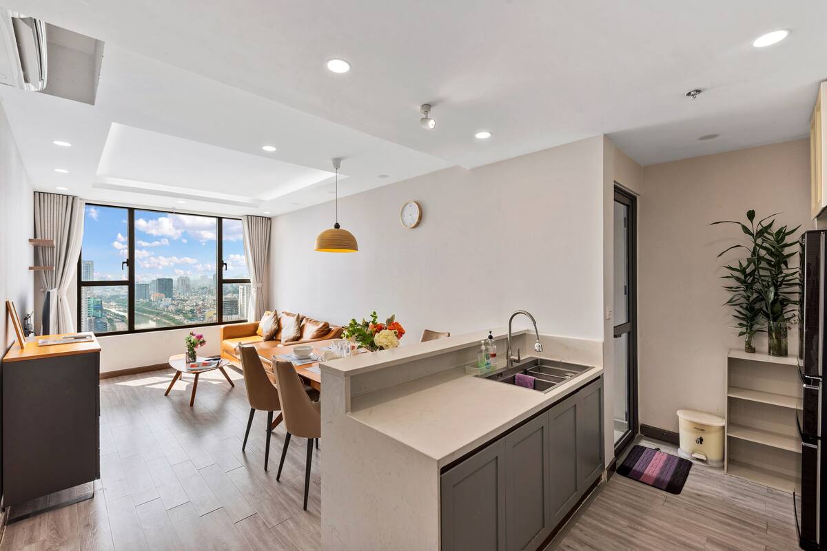 A modern kitchen with a central island, a dining area with a sofa and chairs, and a large window with a city view.