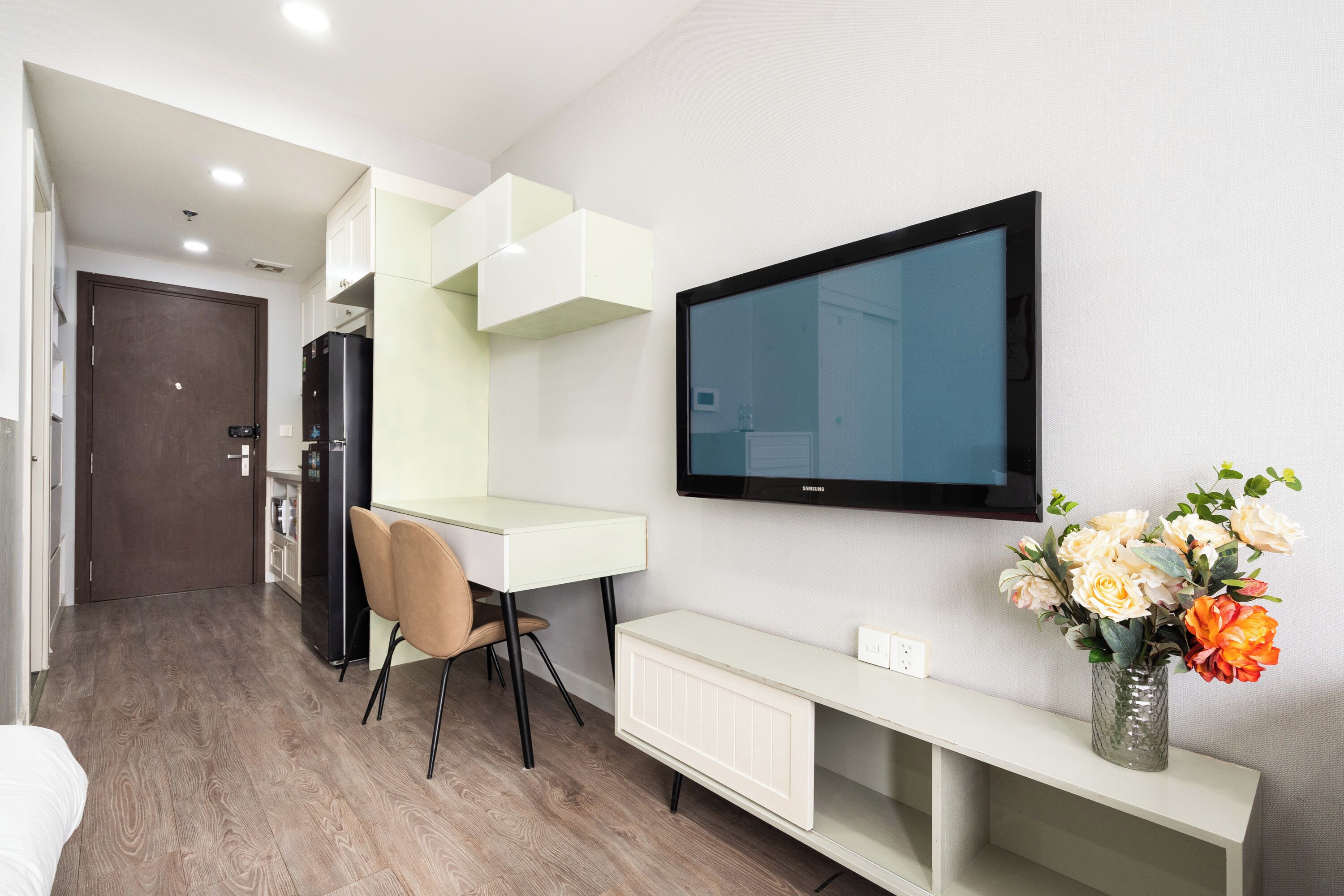A modern hotel room with a wooden floor, a white desk with a chair, a flat-screen TV mounted on the wall, and a vase with flowers on a white console table.