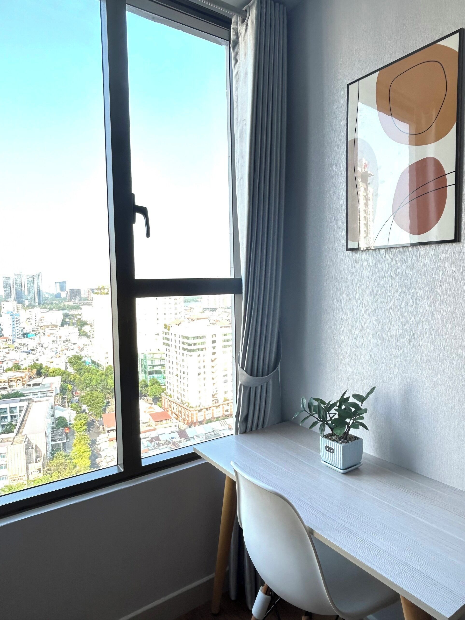 Ein Zimmer mit Schreibtisch, einer Topfpflanze, einem Vorhangfenster und einem gerahmten Bild an der Wand.
