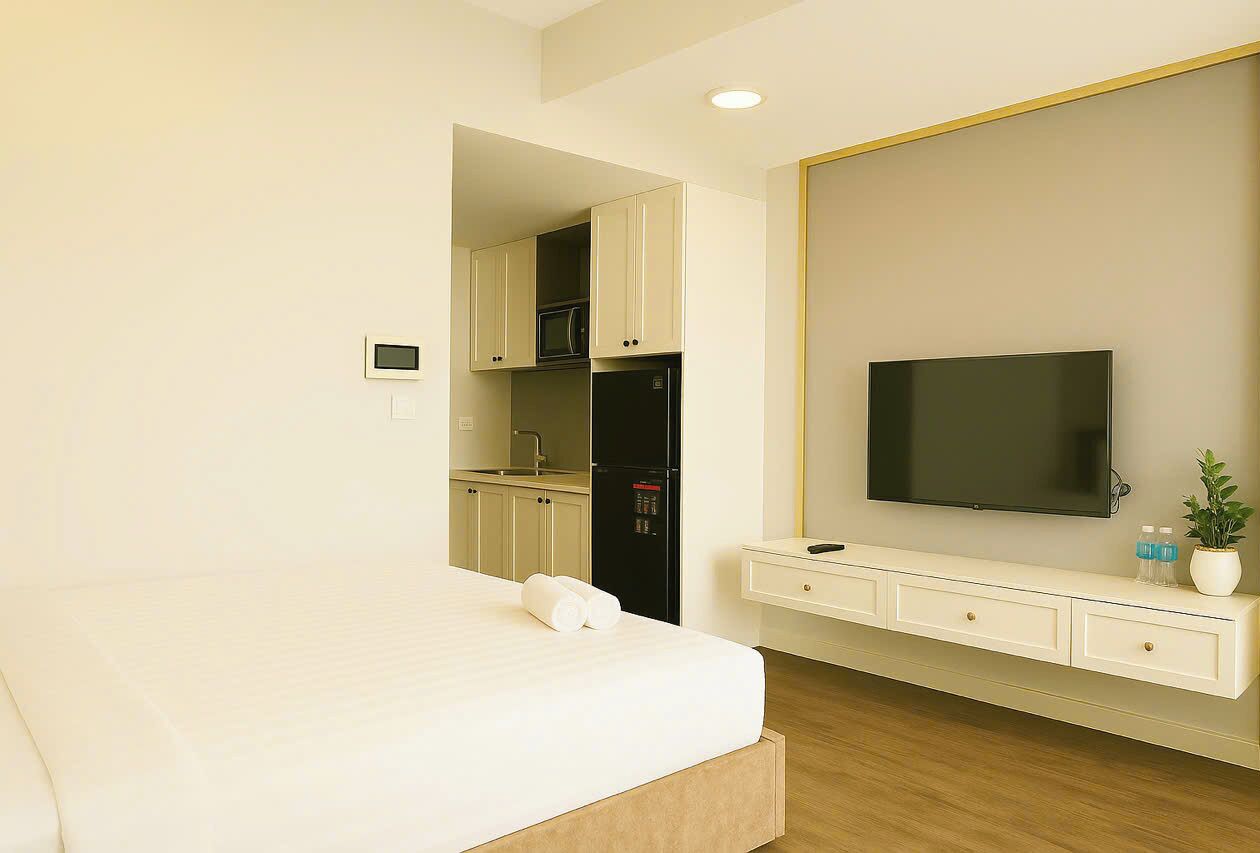 Habitación de hotel con cama, televisor montado en la pared, pequeña cocina y una mesita consola blanca.