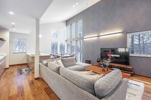 A modern living room with a large gray sofa, wooden flooring, a flat-screen TV, and a dining area with a table and chairs.