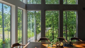 Un comedor con una mesa grande y sillas, amplias ventanas que ofrecen vista a un área boscosa.