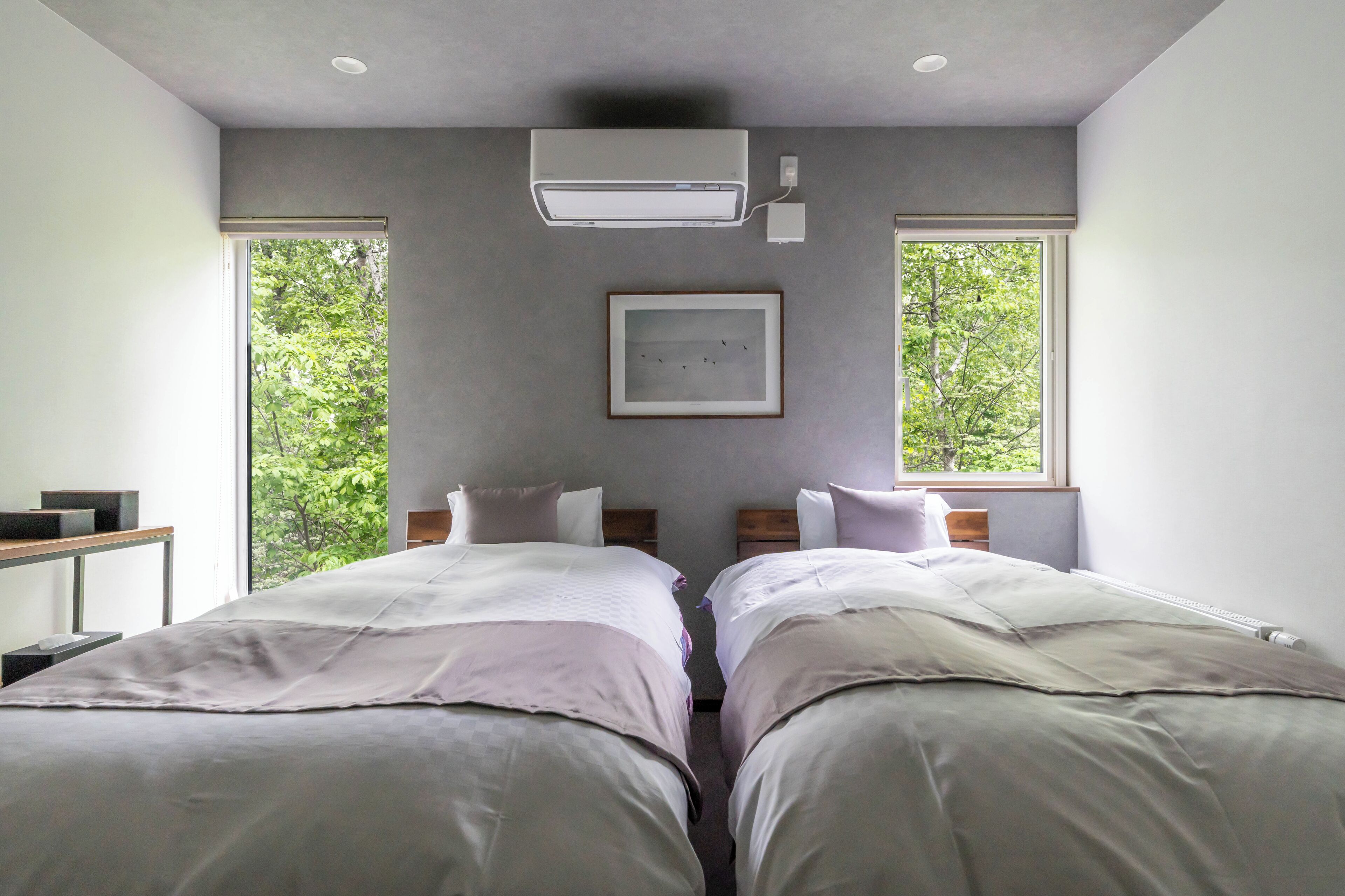 A modern hotel room with two beds, a ceiling air conditioner, a framed picture, and large windows with a view of trees.