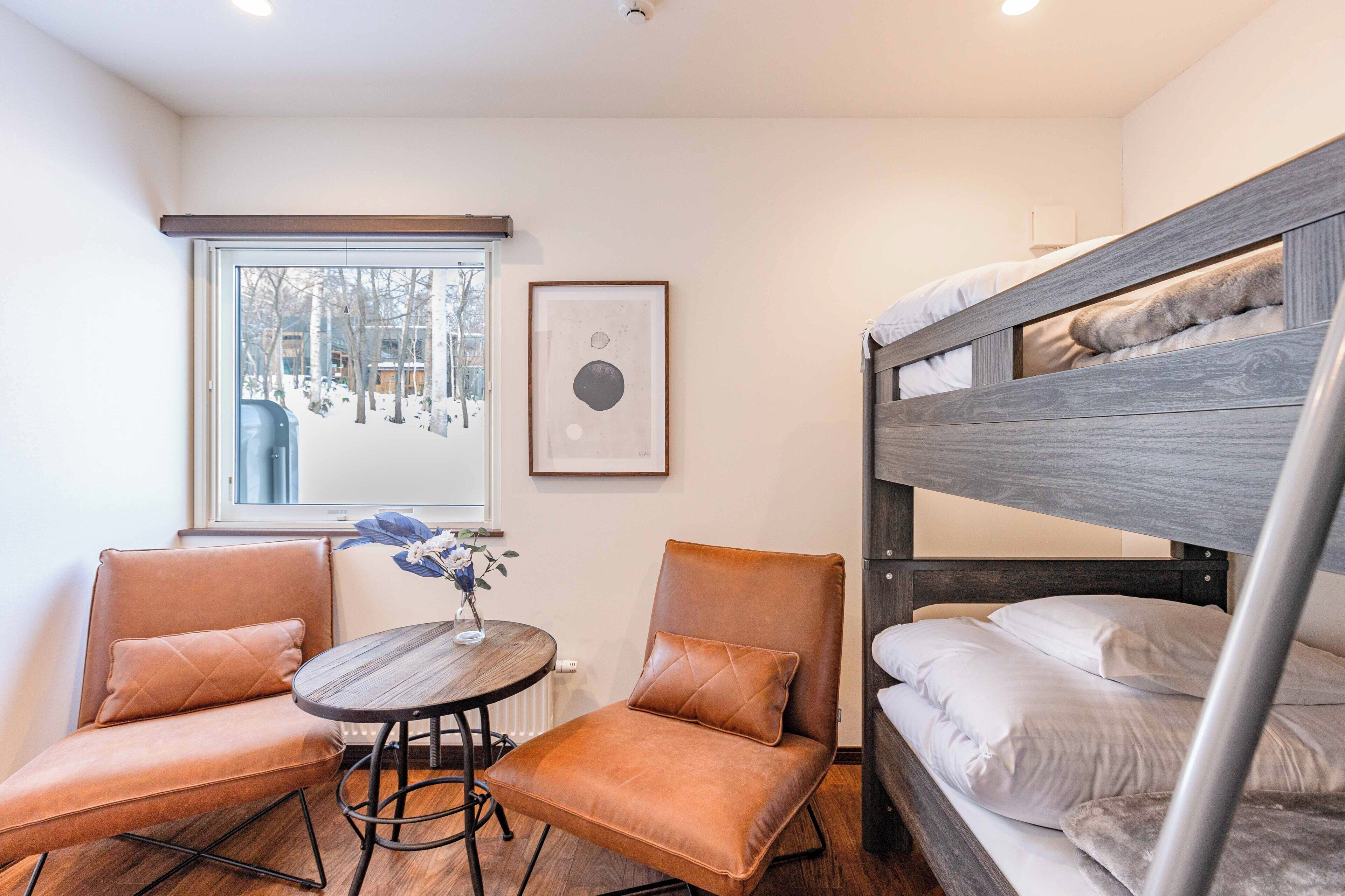 A room with a bunk bed, two leather armchairs, a small round table with a vase, and a window with a view of trees.