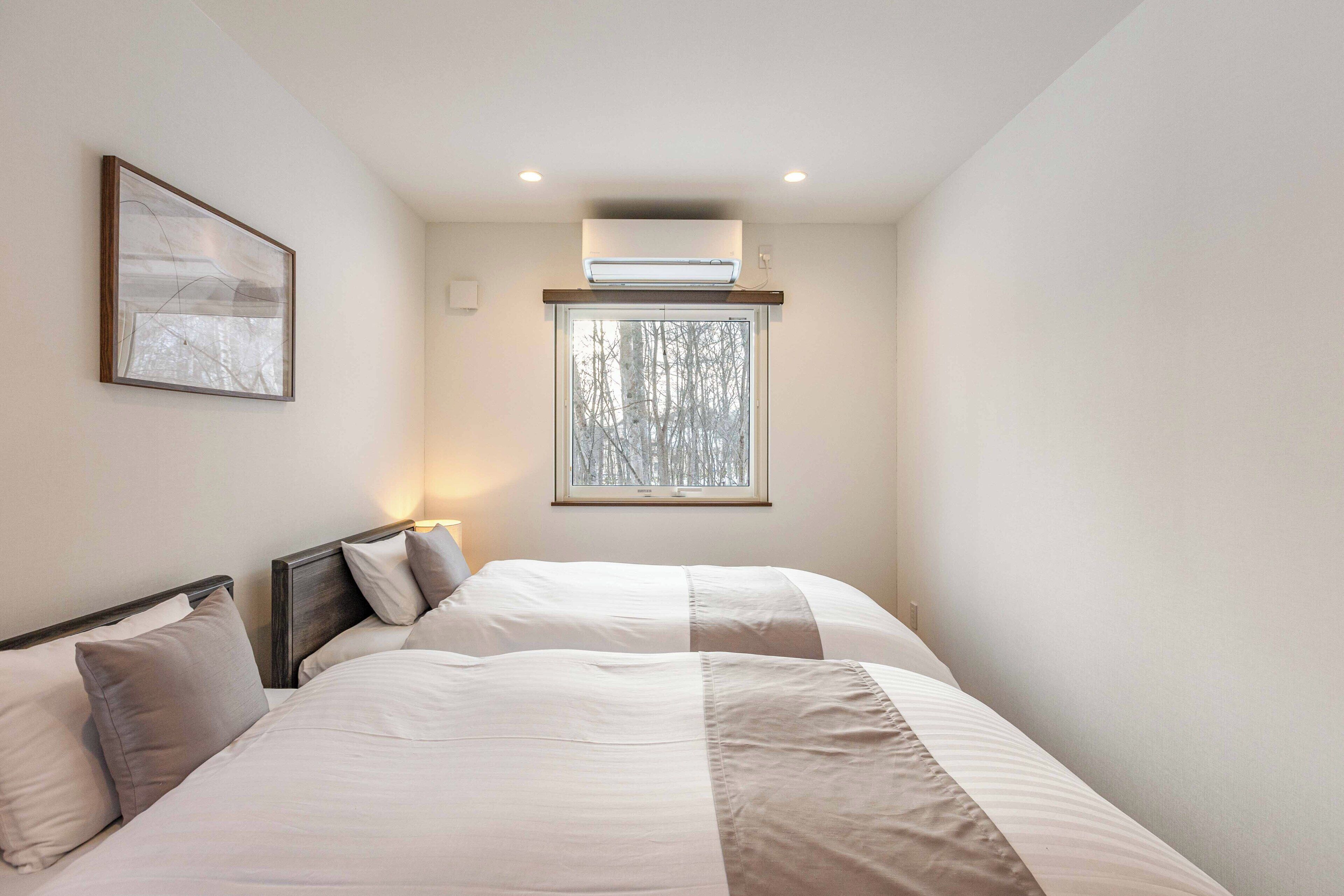 Un dormitorio con dos camas, una ventana con vistas a árboles, un equipo de aire acondicionado y un cuadro en la pared.