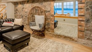 Un salon chaleureux avec un mur en pierre, un grand miroir qui reflète la vue sur la mer, un fauteuil en osier et un canapé en cuir agrémenté de coussins.