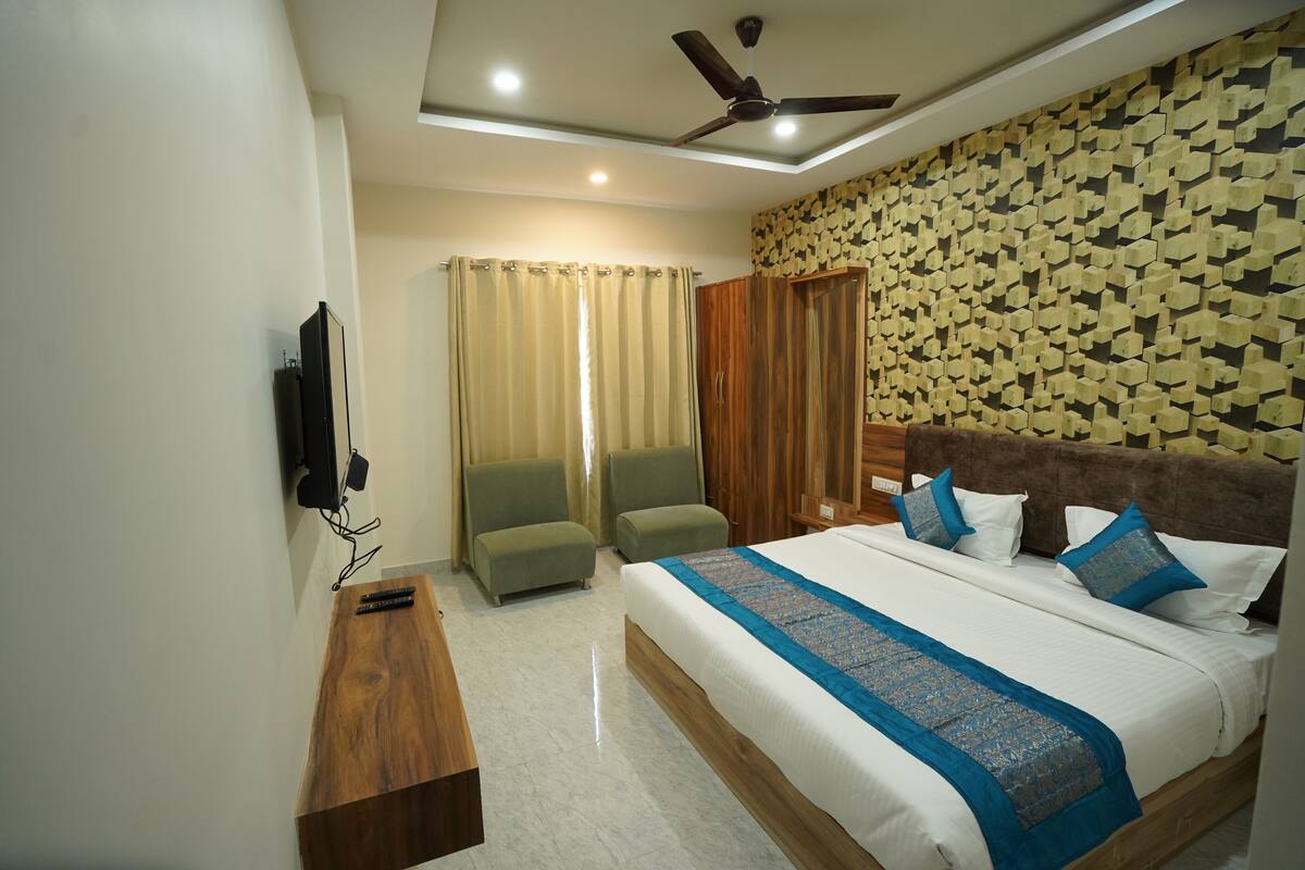 A hotel room with a bed, two armchairs, a wooden bench, a ceiling fan, and a TV mounted on the wall.