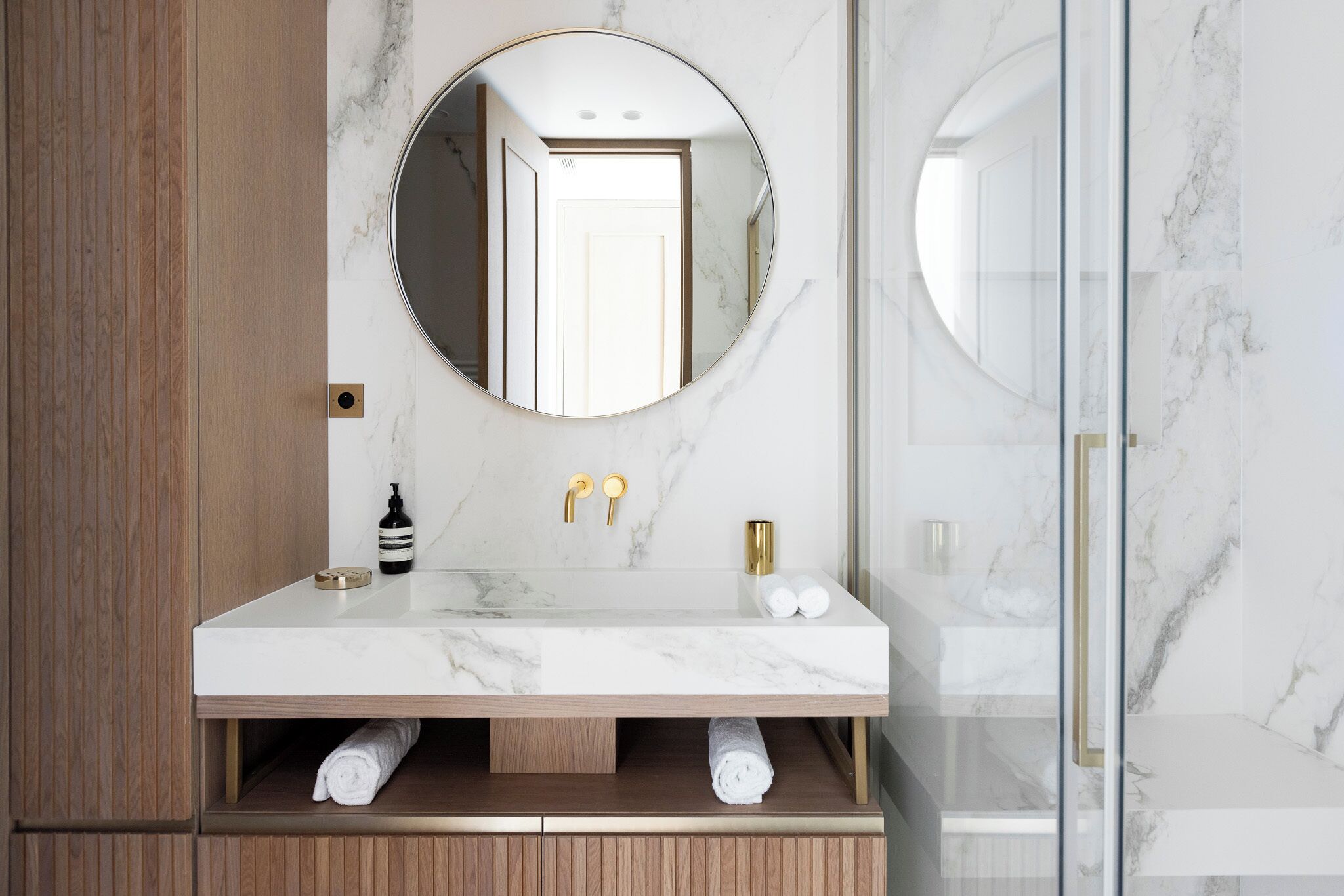 A bathroom with a marble vanity, a round mirror, and a glass shower enclosure.