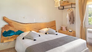 A neatly made bed with a wooden headboard, a bedside table with a lamp, and a view of the outdoors through a window. - Penny Luxury Apartments (Samos)