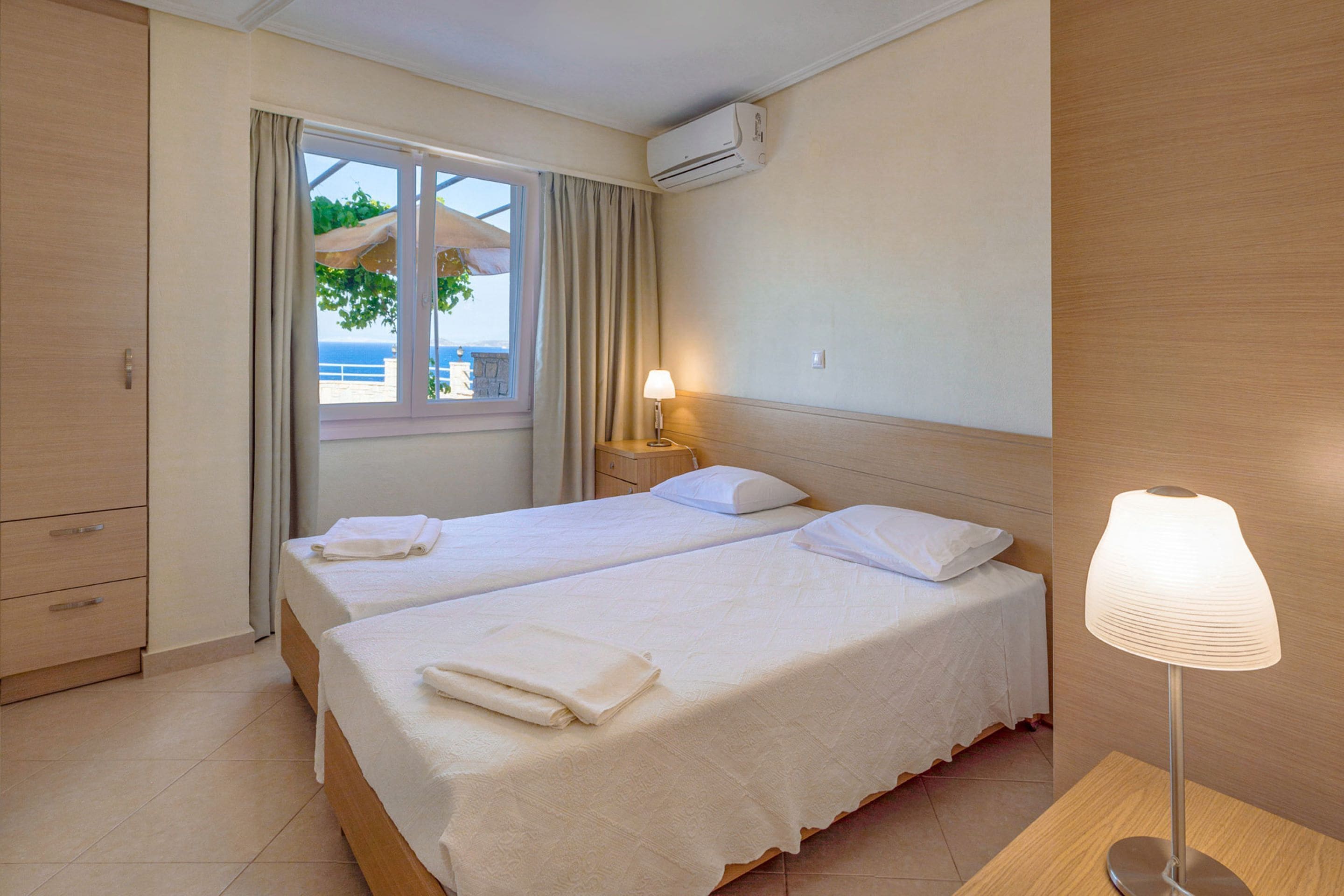 A hotel room with two beds, a window with curtains, an air conditioning unit, a bedside lamp, and a wooden nightstand.