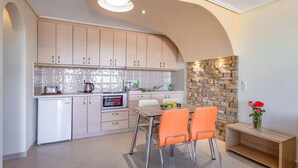 A modern kitchen with light wood cabinets, a built-in oven, and a stone accent wall. There is a dining table with two orange chairs and a vase with flowers on a wooden side table. - Penny Luxury Apartments (Samos)