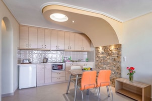 A modern kitchen with light wood cabinets, a built-in oven, and a stone accent wall. There is a dining table with two orange chairs and a vase with flowers on a wooden side table.