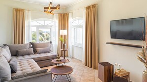 A modern living room with a sectional sofa, two round coffee tables, a flat-screen TV mounted on the wall, and large windows with curtains. - Penny Luxury Apartments (Samos)