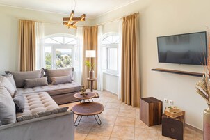 A modern living room with a sectional sofa, two round coffee tables, a flat-screen TV mounted on the wall, and large windows with curtains.