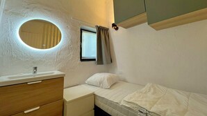 Een kleine, eenvoudige kamer met een enkel bed, een nachtkastje, een spiegel en een raam met gordijnen.