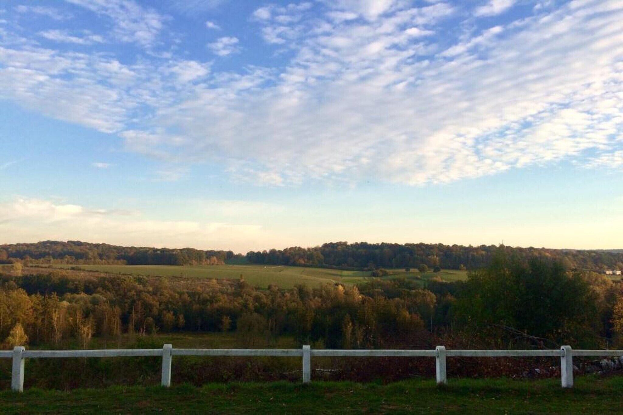 A rural landscape with a white fence in the foreground and a view of rolling hills and trees under a partly cloudy sky.