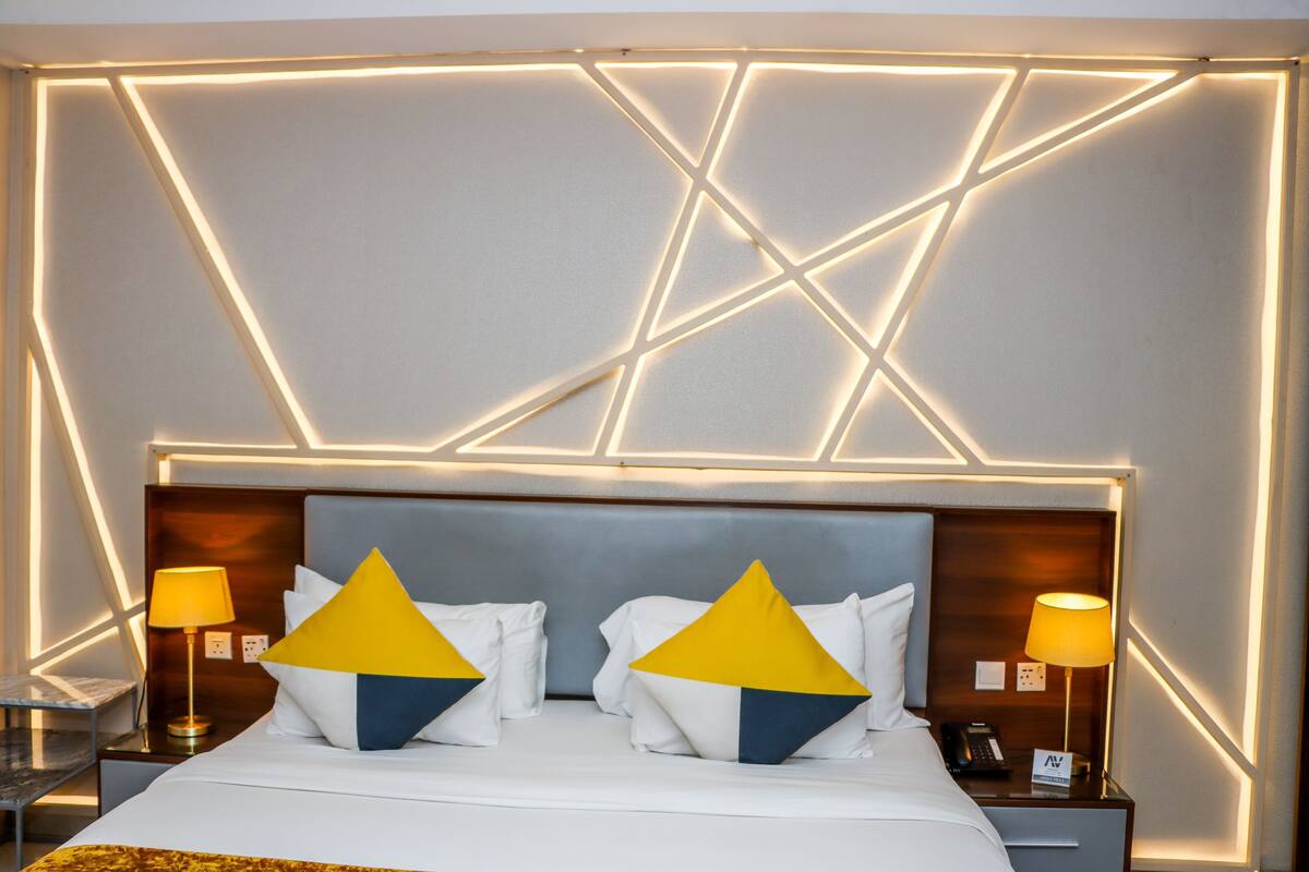 A hotel room with a modern design, featuring a geometric light pattern on the ceiling, a neatly made bed with white bedding and decorative pillows, and bedside tables with lamps.