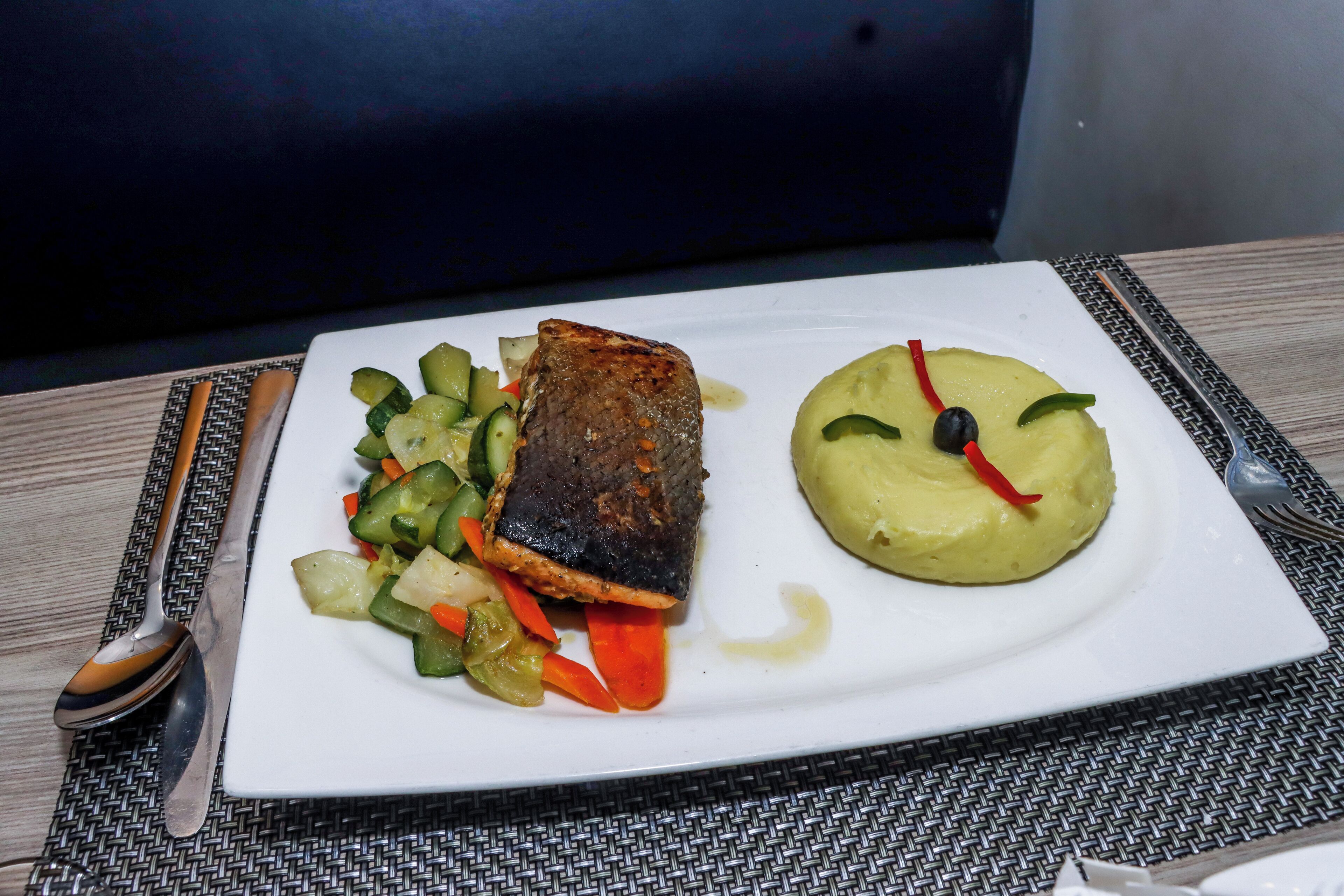 Een wit bord met een stuk gegrilde vis, een hoopje puree en een portie gemengd groentewortels.