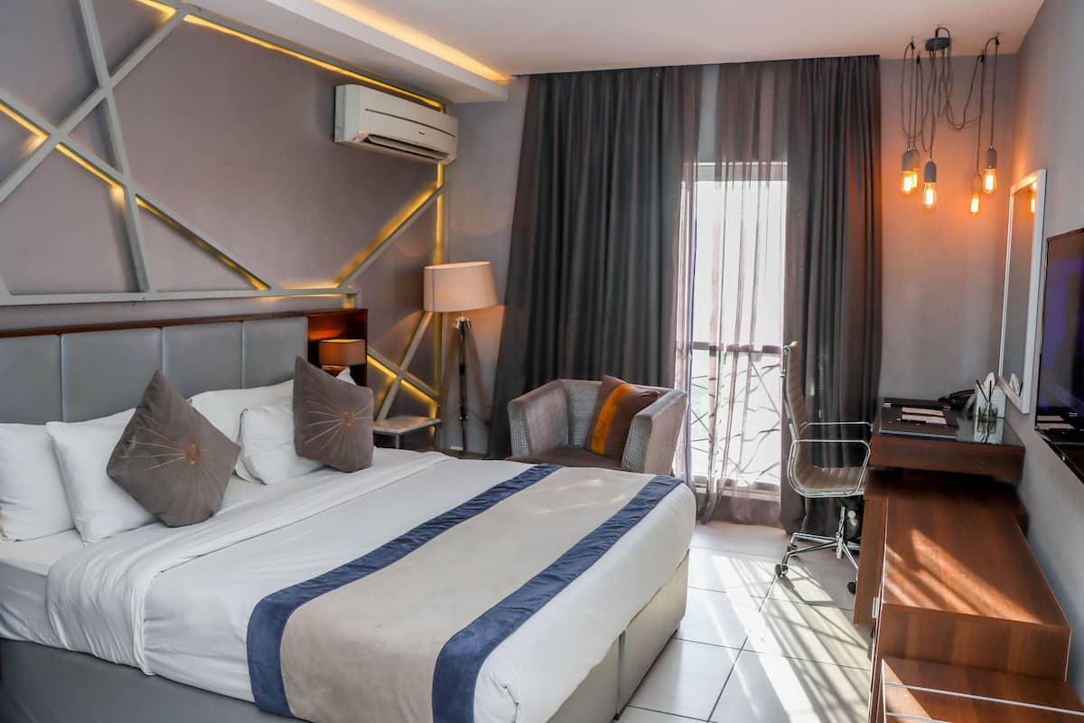 A modern hotel room with a large bed, a desk, and a chair. There is a window with curtains, a lamp, and a TV.
