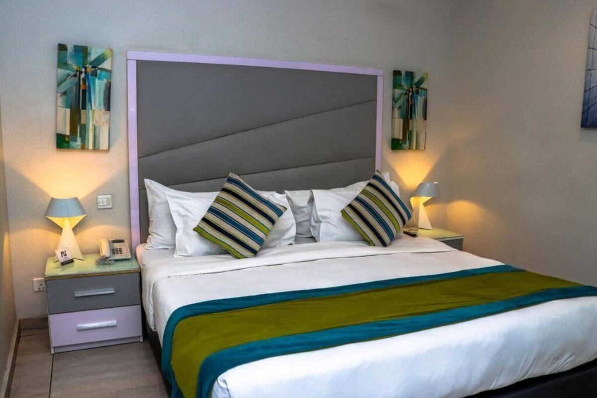 A neatly made bed with striped pillows, a grey headboard, and a teal and green throw blanket. There are two bedside lamps and a small bedside table with a phone and a lamp.