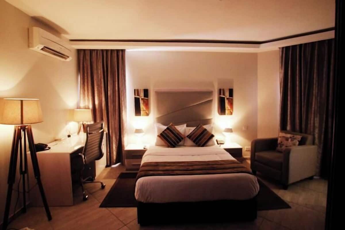 A hotel room with a large bed, bedside tables, a desk, a chair, and an air conditioning unit.