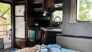 A compact kitchen with a microwave, stove, and sink. There is a dining table with a woven basket and a decorative plate. - Paradise on the Wheels Clearwater Beaches (Largo)