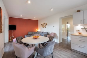 A modern living room with a round table, grey chairs, a red accent wall, a flat-screen TV, and a sofa.