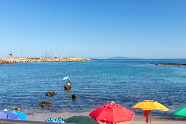 A beach with clear blue water, a dolphin sculpture, and colorful umbrellas.