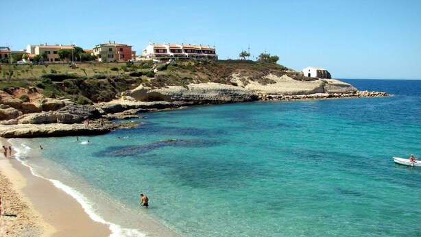 Una playa costera con agua cristalina, costa rocosa y edificios al fondo.