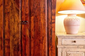 A wooden wardrobe with a brass handle, a white dresser with a lamp, and a beige wall.