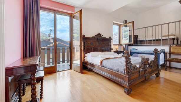 A bedroom with a wooden bed, a desk, a chair, a large window with a view, and a sliding door leading to an outdoor area.